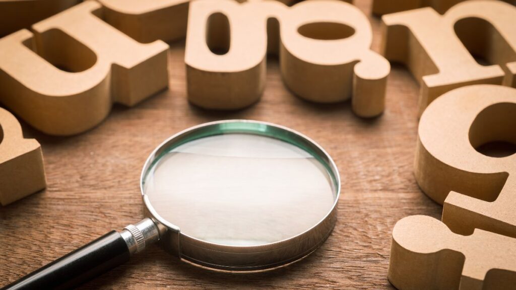 A magnifying glass laying on a wooden surface surrounded by wooden letters.