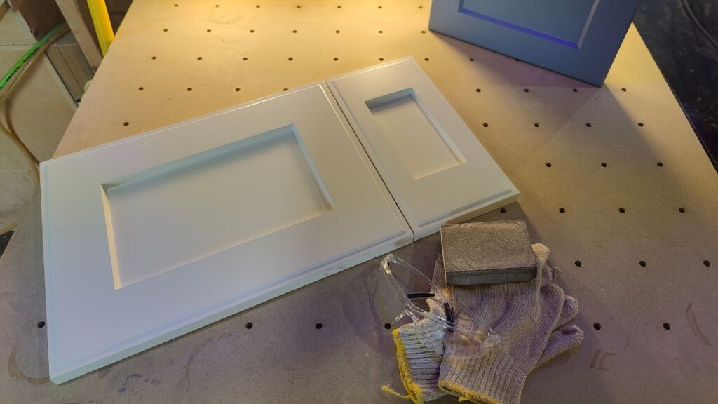 White and blue painted cabinet doors on a pegboard table. Safety glasses, a sanding block and gloves lie in front of the white door and the blue door is visible in the background.