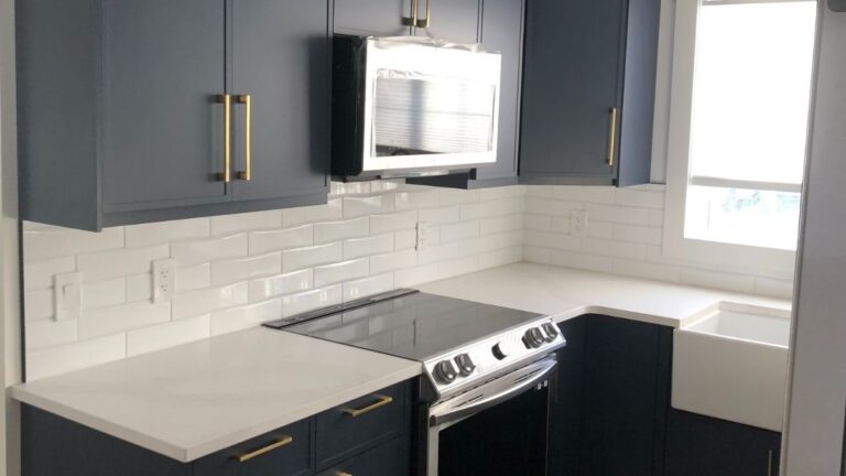 A kitchen with blue painted 1-piece MDF slim shaker cabinet doors, white backsplash and countertops, and brass hardware. Adding finishing or custom details to your order will increase the price.